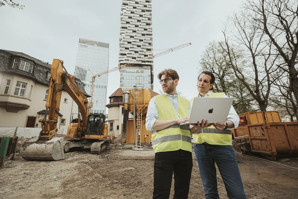 Zwei Personen in Warnwesten arbeiten auf einer Baustelle an einem Laptop.
